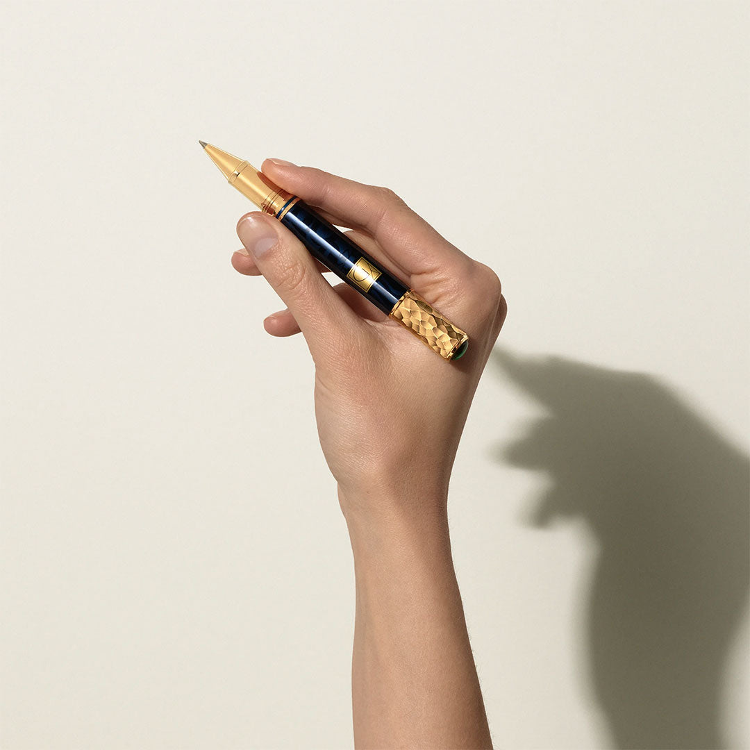 A hand holds the Montblanc Masters of Art Homage To Gustav Klimt Limited Edition 4810 Rollerball Pen, its gold and dark blue design reflecting Vienna Secession style, casting a refined shadow on a plain light background.