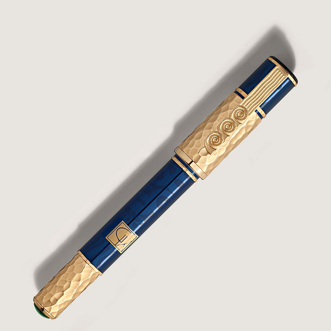The Montblanc Masters of Art Homage To Gustav Klimt Limited Edition 4810 Rollerball Pen features a dark blue body, gold textured spiral accents inspired by the Vienna Secession, and a square emblem, displayed on a light surface with a soft shadow.