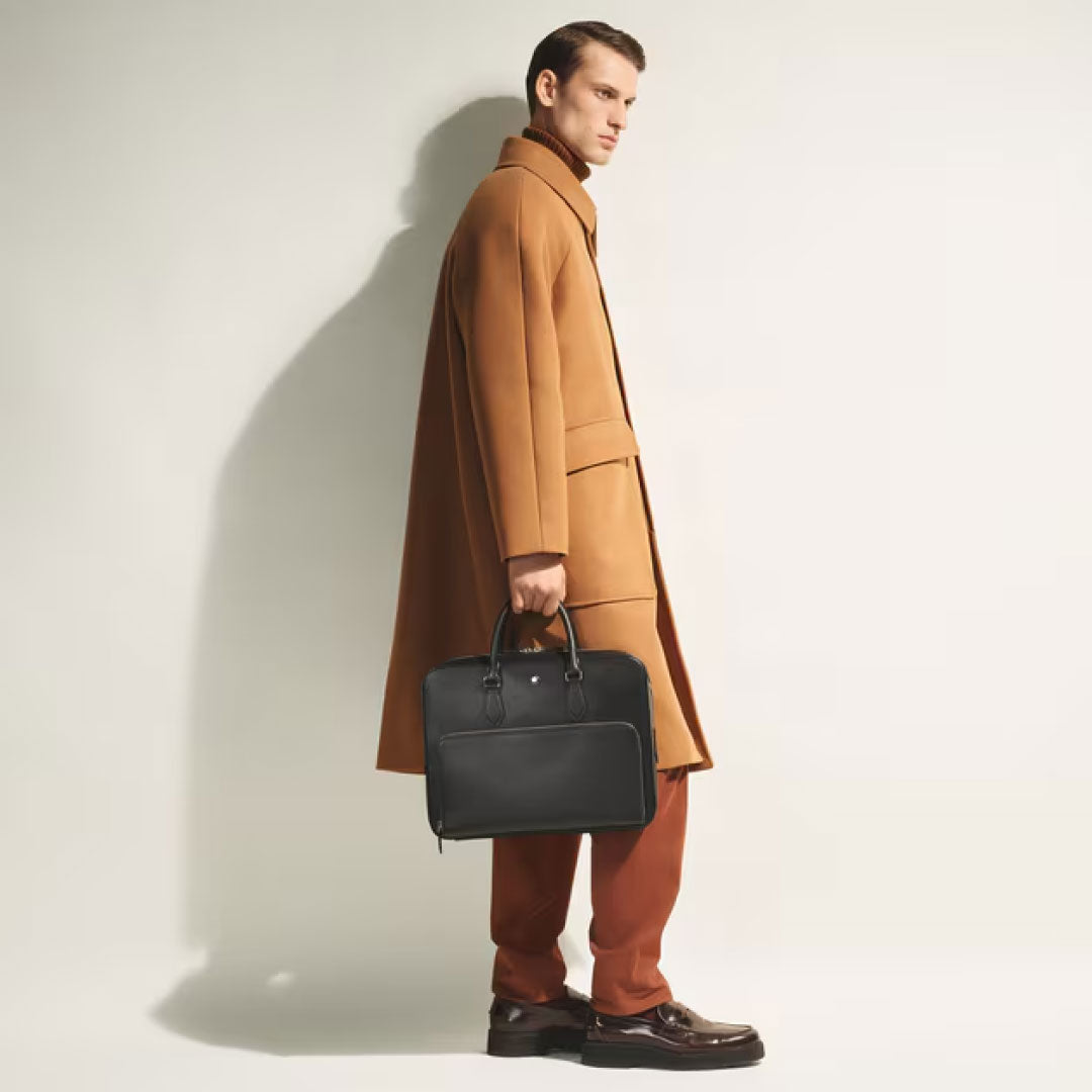 A man in a tan overcoat and rust pants stands sideways against a plain background, holding a Montblanc Sartorial Medium Leather Document Case Bag with Pocket by his side and wearing dark dress shoes.