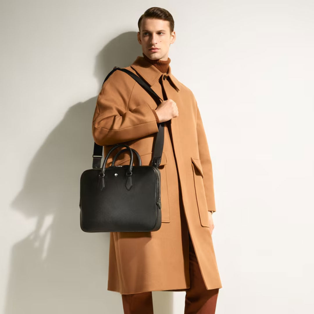 A man in a camel overcoat and brown pants stands against a light background, carrying the Montblanc Sartorial Medium Leather Document Case Bag with Pocket by Montblanc.