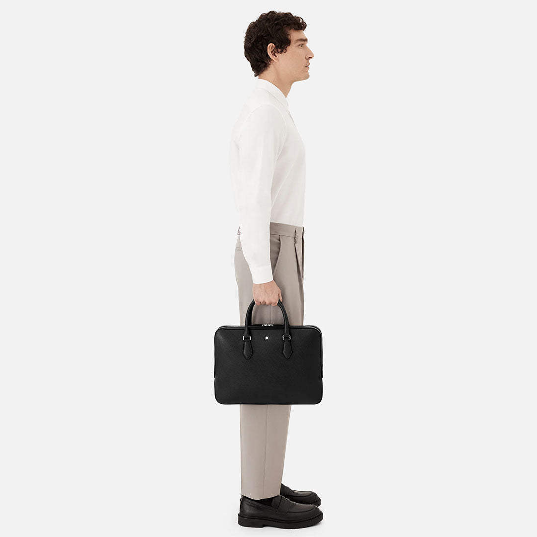 A man in a white shirt and beige pants stands in profile, holding the Montblanc Sartorial Thin Leather Document Case Bag by his side against a plain white background.