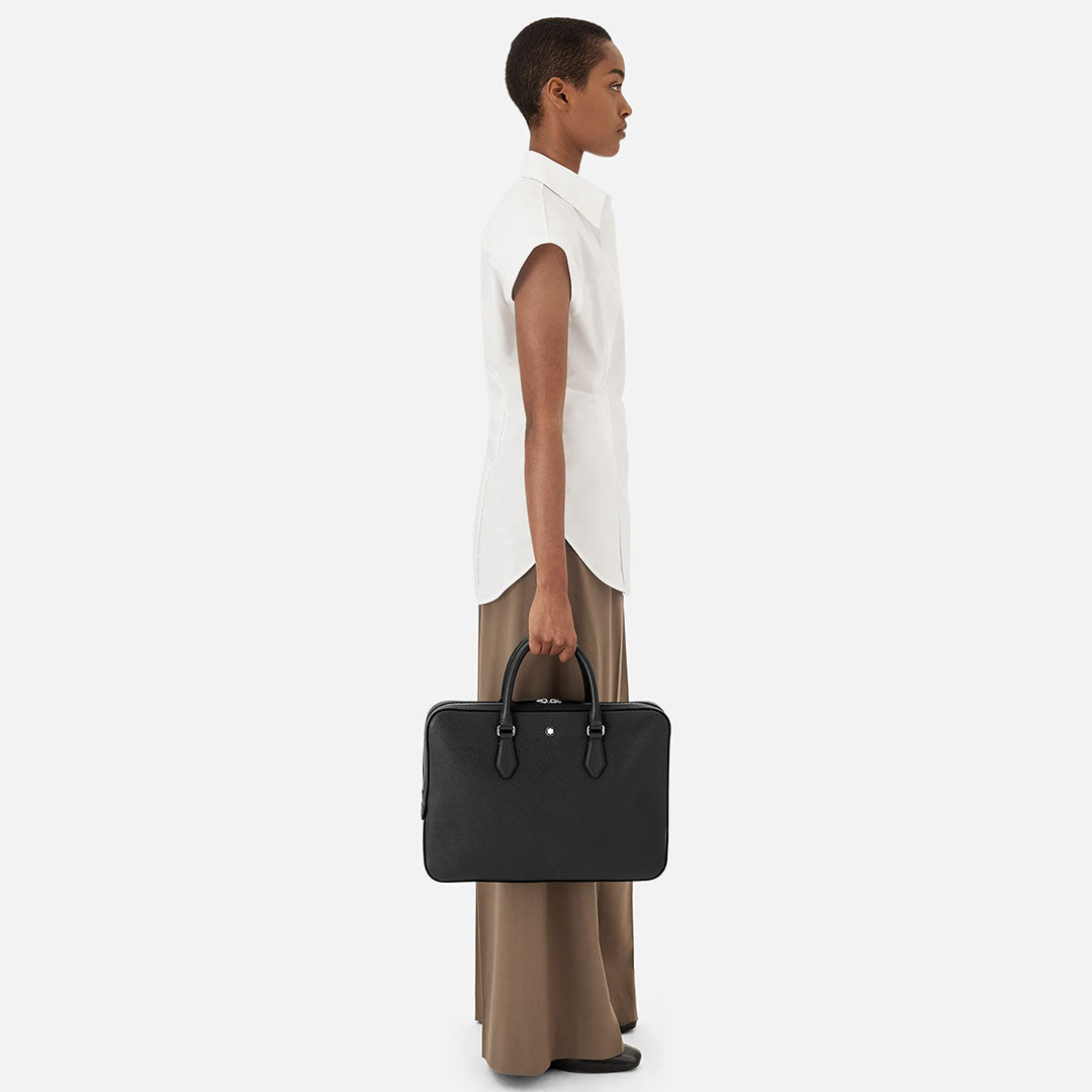 A person with short hair, dressed in a white sleeveless blouse and taupe wide-leg pants, stands in profile holding the Montblanc Sartorial Thin Leather Document Case Bag by their side against a plain white background.
