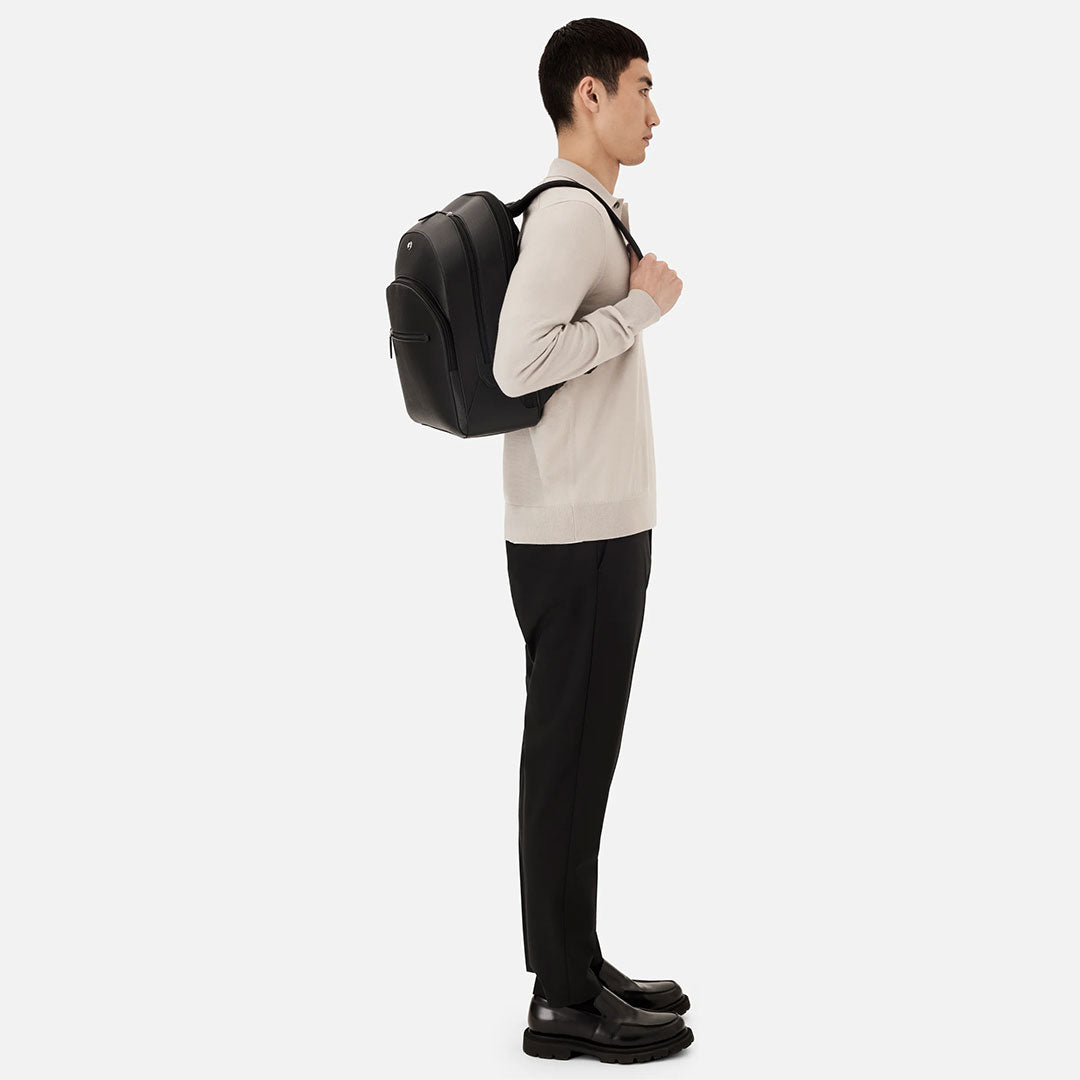 A man in profile against a plain background wears a light sweater, black pants and shoes, carrying the Montblanc Sartorial Large 3 Compartments Leather Backpack—an ideal choice from Montblanc for business trips.