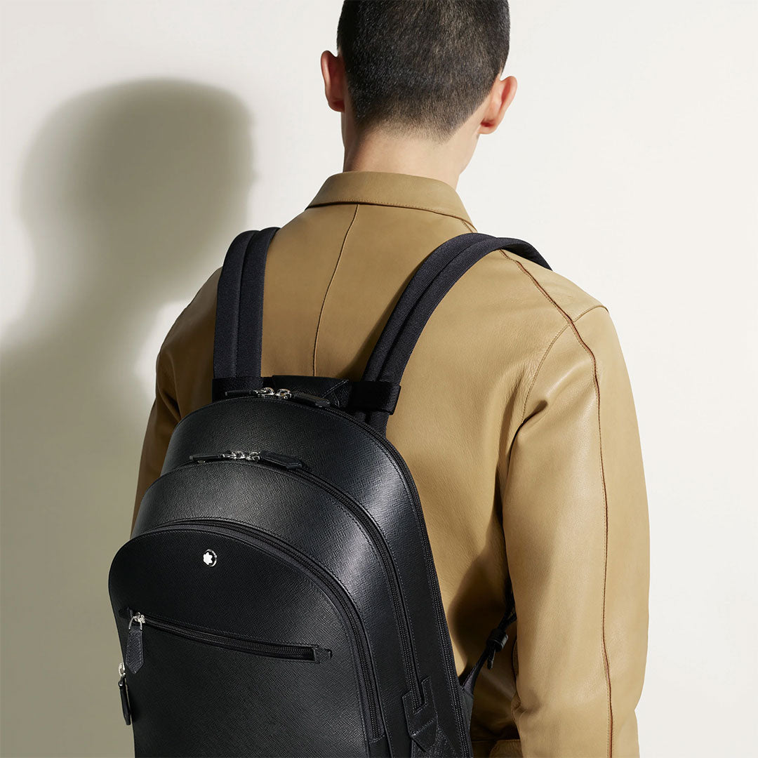 A person in a tan jacket is seen from behind carrying a Montblanc Sartorial Medium 3 Compartments Leather Backpack Bag, featuring a small white emblem on the top pocket. The background is plain and well lit.