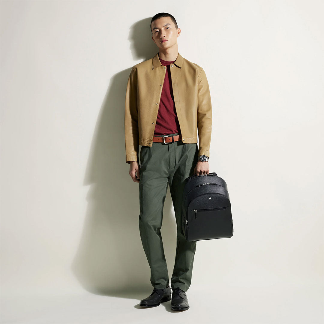 A man stands against a white background wearing a tan jacket, red shirt, green pants, black shoes, and holding a Montblanc Sartorial Medium 3 Compartments Leather Backpack Bag in his left hand.