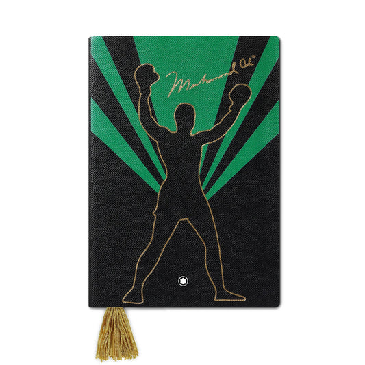 The Montblanc Muhammad Ali #146 Notebook features a boxer with raised gloves silhouetted against a green and black sunburst backdrop, with "Muhammad Ali" elegantly printed in gold at the top. A gold tassel bookmark hangs below this tribute to one of history's great figures.