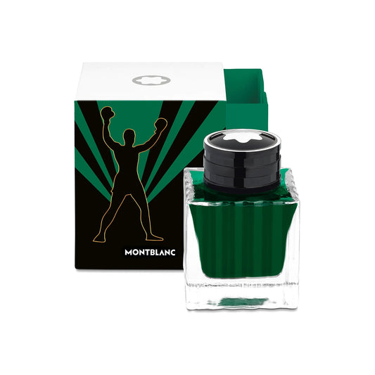 A Montblanc Muhammad Ali Ink Bottle rests in front of a partially open box, which features a silhouette of a person with raised arms against green rays and the "Montblanc" brand name. This design is from the Great Characters series, celebrating icons such as Muhammad Ali.