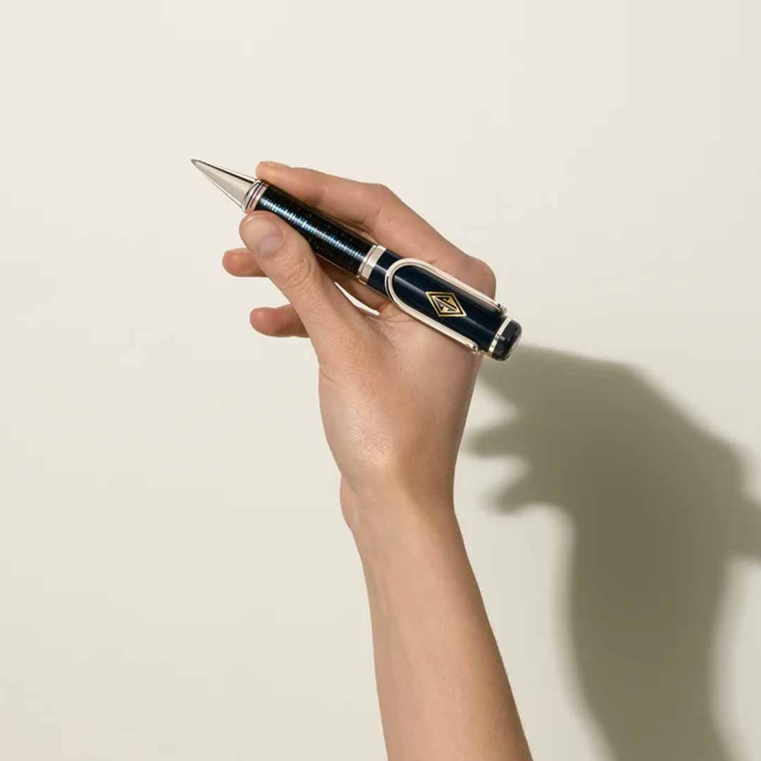 A hand elegantly holds the Montblanc Great Characters Homage to The Great Gatsby Special Edition Ballpoint Pen, featuring a black and silver design with a decorative emblem that evokes the glamour of the Jazz Age, against a plain light background. The shadow cast by the hand vividly reflects the sophistication of The Great Gatsby era.