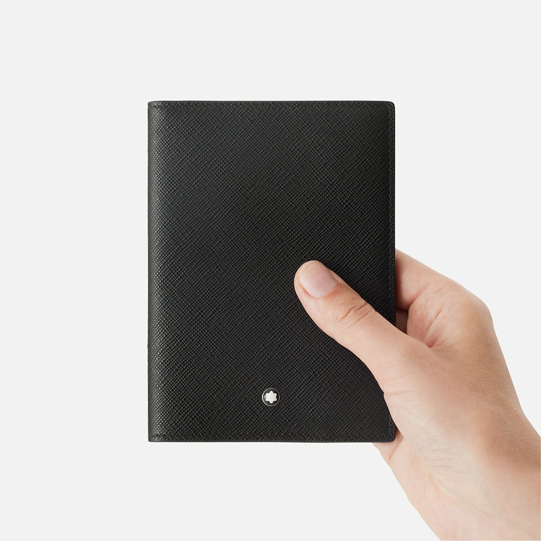 A hand holds a closed Montblanc Sartorial Leather Passport Holder in black, featuring a small round white Montblanc emblem near the bottom center, set against a plain white background.