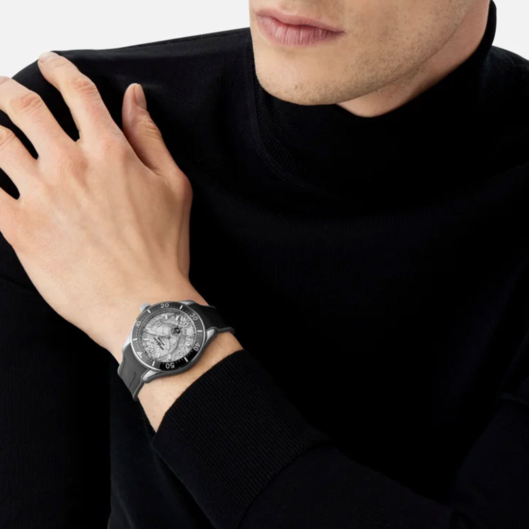 A person in a black turtleneck sweater showcases the Montblanc 1858 Iced Sea Automatic Date 41mm Watch, featuring an intricately detailed face and dark strap. The watch's automatic date function is prominently displayed on their left wrist as they gently touch their shoulder with their right hand.