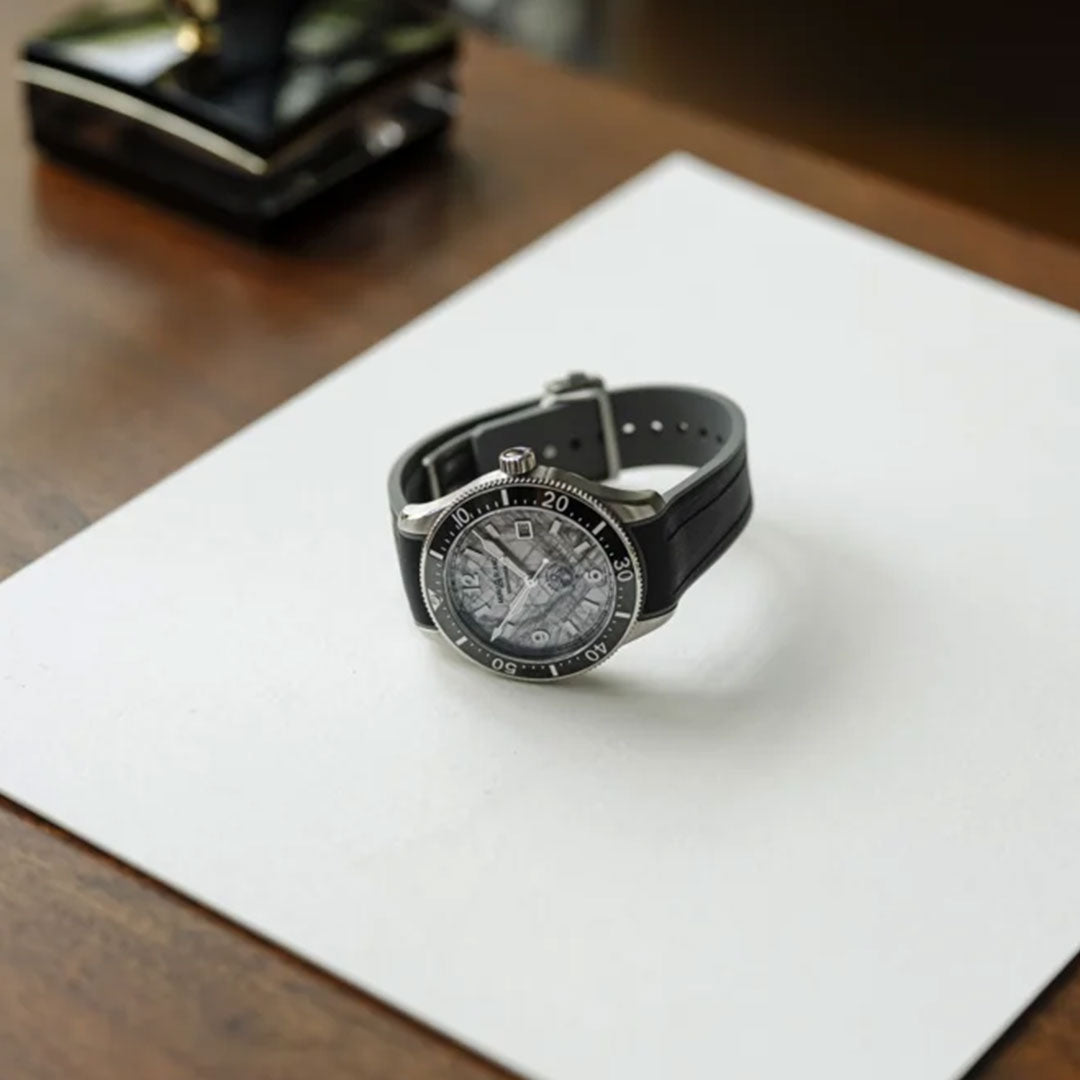 A Montblanc 1858 Iced Sea Automatic Date 41mm Watch with a black strap rests on a white sheet of paper atop a wooden surface. This scuba diving watch showcases a detailed dial adorned with silver markers and hands, and its allure is enhanced by an automatic date function. In the background, you can see a pen stand.