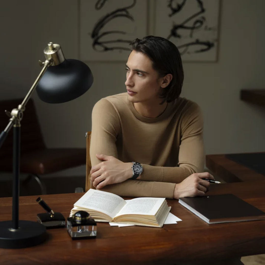 A person with long hair sits at a wooden desk, glancing to the side. They wear a beige sweater and a Montblanc 1858 Iced Sea Automatic Date 41mm Watch. An open book, notebook, and lamp adorn the desk while abstract art decorates the wall in the background.