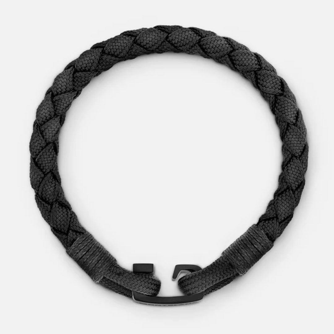 The Montblanc T-Hook Nylon Bracelet, featuring a braided rope design in black with a sleek black clasp and black PVD coating, is elegantly displayed in a circle on a light gray background. This bracelet combines simplicity and elegance with its woven texture and T-Hook closing.
