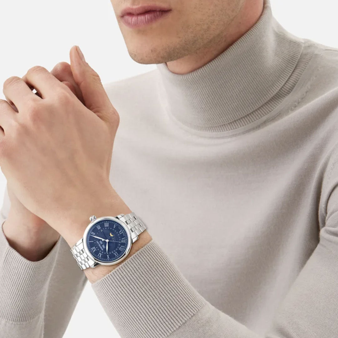 A person wearing a light gray turtleneck sweater and clasping their hands together showcases the Montblanc Star Legacy Full Calendar Automatic 42mm Watch. This silver timepiece features an intricate blue face and comes equipped with an interchangeable strap system, set against a plain white background.