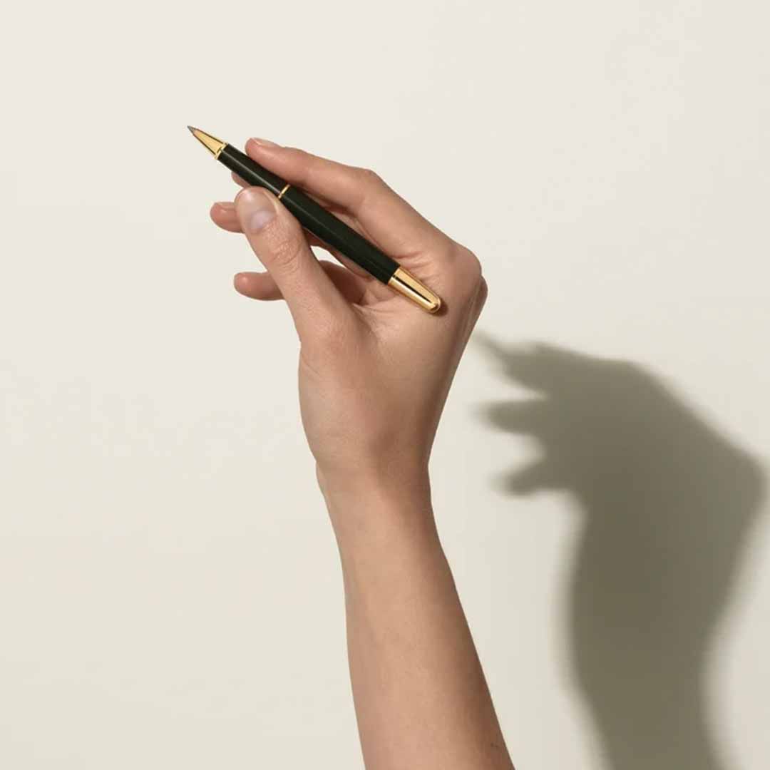 A hand holding a Montblanc Meisterstuck The Origin Collection Doue Classique Rollerball Pen, with its green and gold design, casts the shadow of a playful animal figure on the wall against a light background, highlighting the elegance of this exquisite writing instrument.