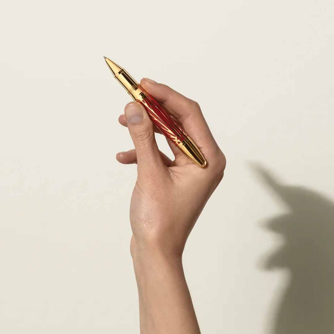 A hand elegantly holds the Montblanc Meisterstück The Origin Collection Solitaire Legrand Rollerball Pen vertically against a plain beige background. Its gold and red design casts a shadow that intriguingly resembles a rabbit, adding an imaginative twist to the timeless craftsmanship of Montblanc's collection.