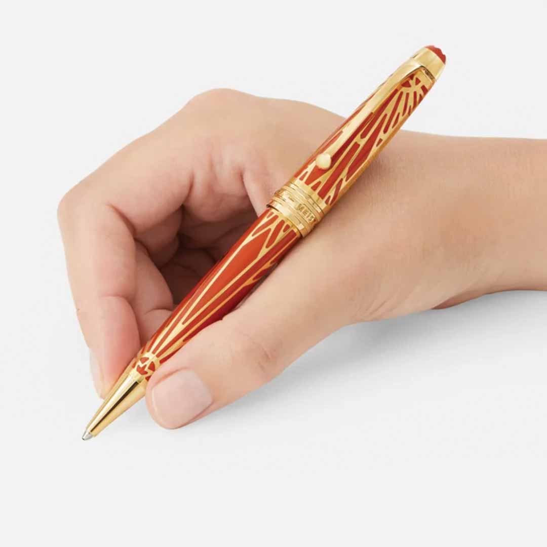 A hand delicately holds a Montblanc Meisterstuck The Origin Collection Solitaire Midsize Ballpoint Pen against a white background. This fine writing instrument, crafted by Montblanc, features intricate golden patterns that enhance its elegant design.