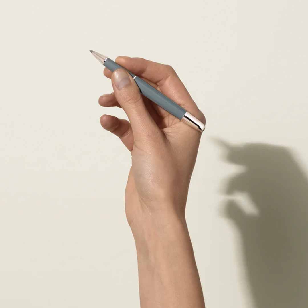 A hand holds a sleek, metallic Montblanc Meisterstück X Olympic Heritage Chamonix 1924 Classique Rollerball Pen against a light wall, casting a distinct shadow resembling a rabbit. The pen's design wonderfully enhances the smooth background, blending elegance and creativity effortlessly.