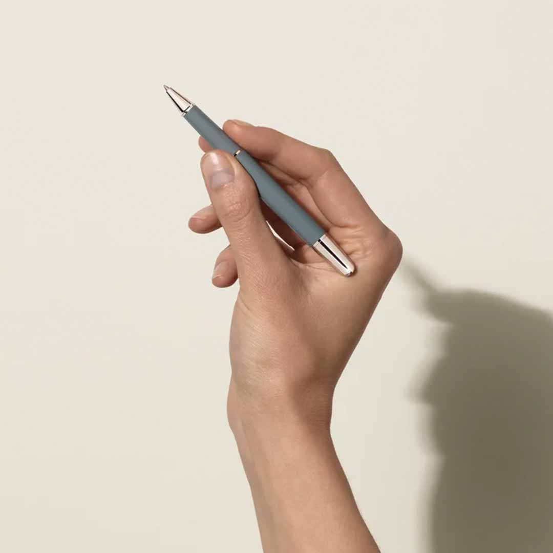 A hand holds a sleek, gray Montblanc Meisterstuck X Olympic Heritage Chamonix 1924 Doue Classique Rollerball Pen against a light beige background. The shadow of the hand and pen is subtly cast on the wall. The image highlights the pen’s minimalist design, reminiscent of Montblanc's timeless elegance.