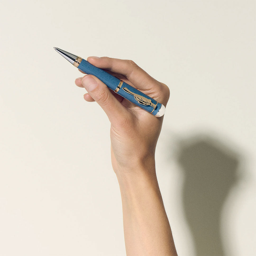 A hand holds the Montblanc Writers Edition Homage to Johann Wolfgang von Goethe Limited Edition Ballpoint Pen, featuring a blue and gold feather design, against a plain light background with a soft shadow.