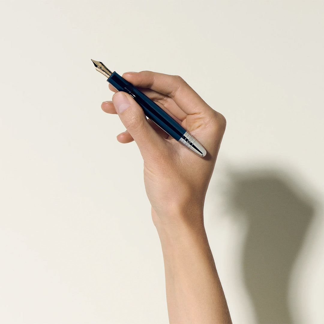 Against a light backdrop, a hand holds a Montblanc Meisterstuck Around the World in 80 Days Legrand Fountain Pen, its shiny blue barrel and gold nib casting a shadow, poised for adventures reminiscent of Jules Verne's tales.