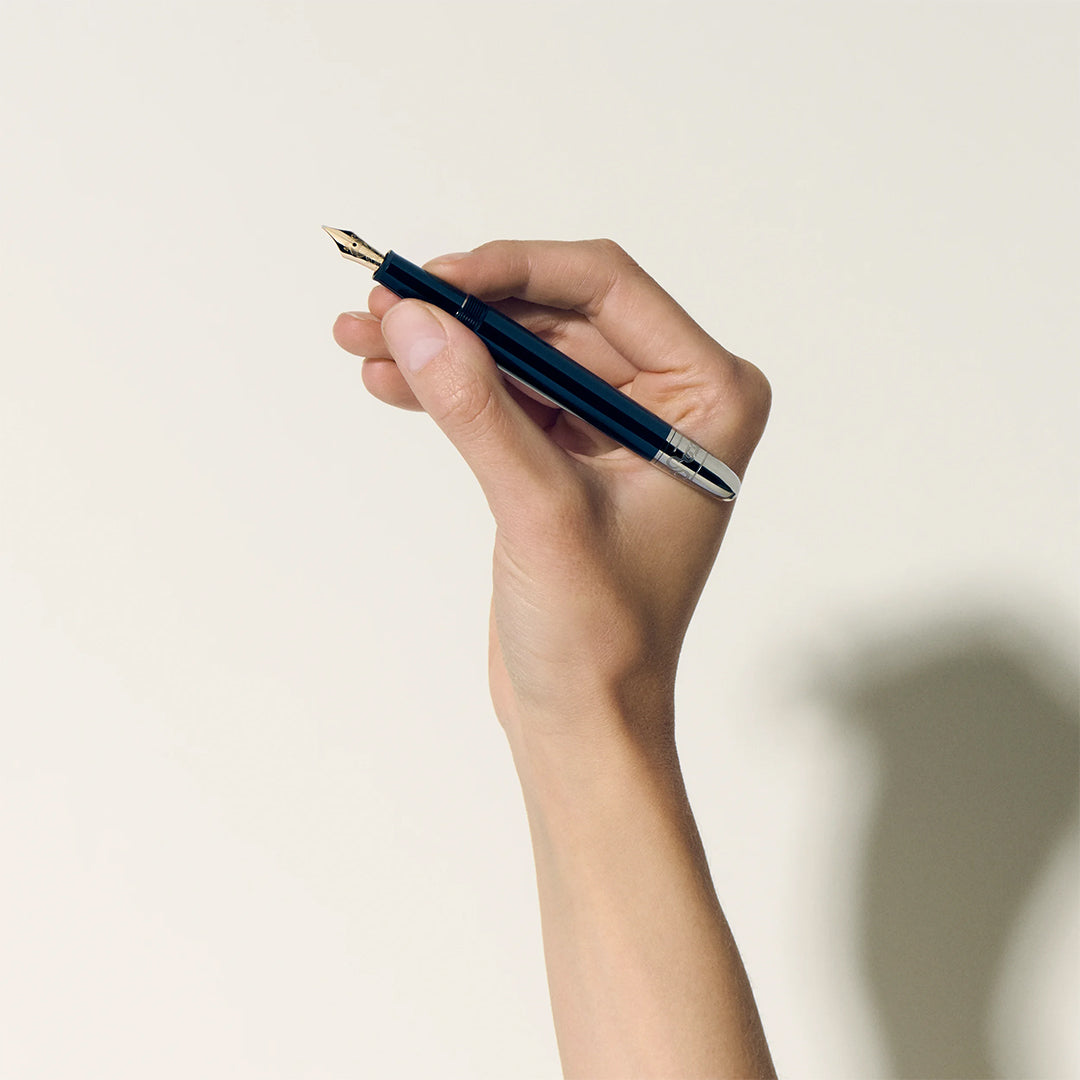 A hand holds a Montblanc Meisterstück Around the World in 80 Days Classique Fountain Pen against an off-white background, casting shadows on the right. The pen is angled upwards, poised as if to write, inspired by Jules Verne's adventurous spirit.