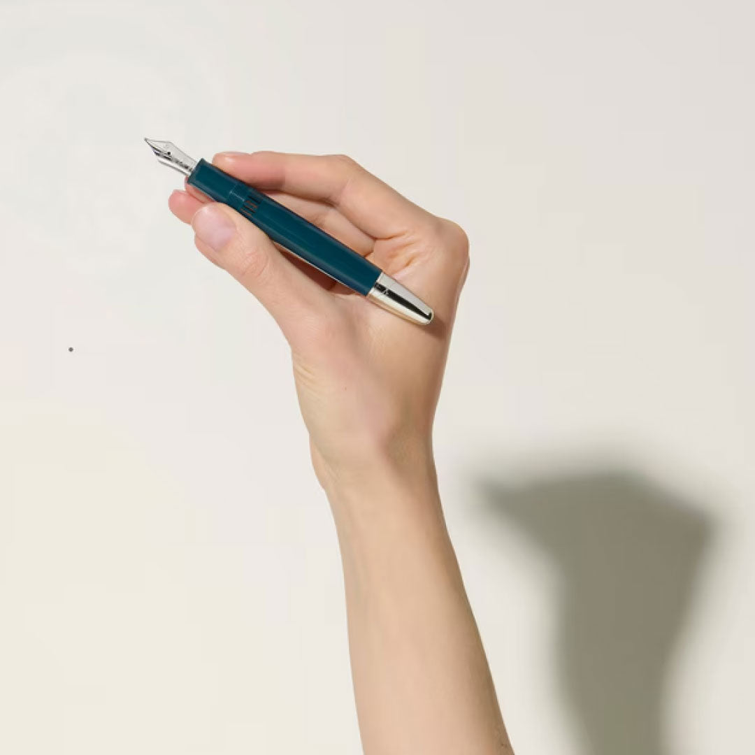 A hand holds a Montblanc Meisterstuck Romeo & Juliet LeGrand Fountain Pen, featuring a silver tip, against a plain light background. The shadow of the hand is visible on the surface.