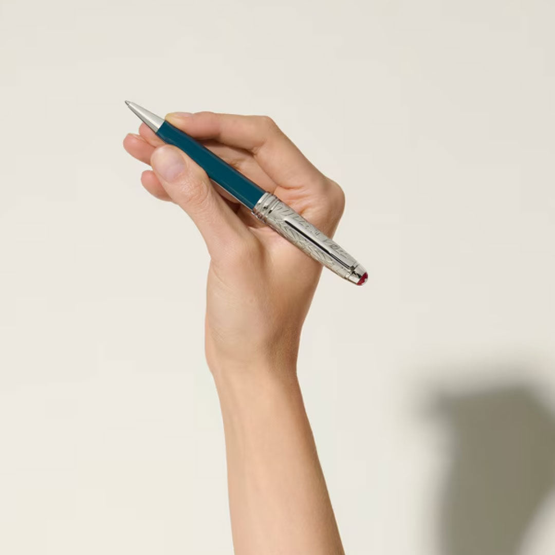A hand holds the Montblanc Meisterstuck Romeo & Juliet Doue Midsize Classique Ballpoint Pen, featuring blue and silver accents, against a light background, its elegant design pointed upward and casting a faint shadow.