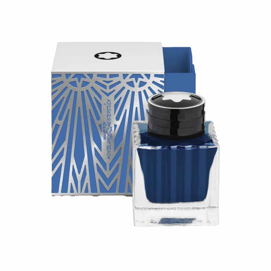 The Montblanc Meisterstuck The Origin Collection Blue - 50ML, a clear glass ink bottle with a black and white cap filled with Origin Blue ink, sits in front of an intricately designed blue and silver box. The box showcases geometric patterns and reflects Montblanc's exquisite style, appearing partially open.
