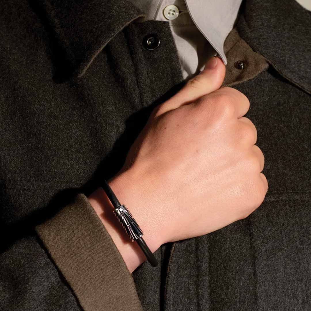 A person wearing a dark green coat holds their collar with one hand, showcasing a minimalist Montblanc Meisterstuck Origins blue leather bracelet on their wrist. The image highlights the silver clasp and distinctive Solitaire pattern of the bracelet against the fabric of the coat.
