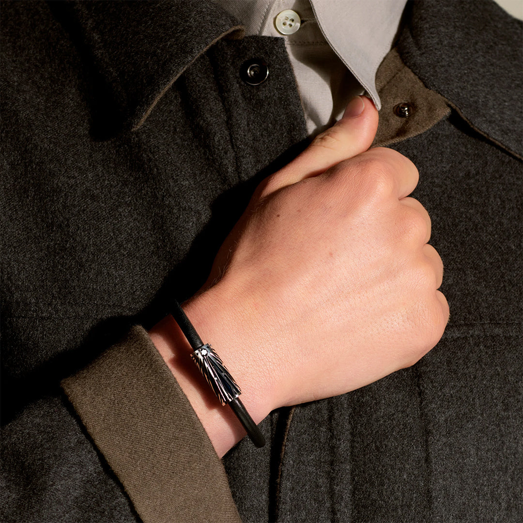 A close-up of a hand with the Montblanc Meisterstuck The Origin Collection Leather Bracelet and small tassel design adds elegance against a dark gray coat. The person hooks their thumb inside the coat, revealing a peek of a buttoned shirt collar underneath.