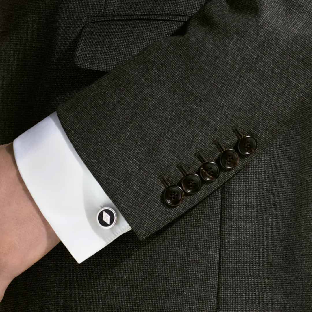A close-up reveals an arm clad in a dark suit jacket, with a white shirt cuff peeking beneath, evoking the era of The Great Gatsby. The cuff is adorned with Montblanc Great Characters Homage to The Great Gatsby Cufflinks, hexagonal and crafted from stainless steel. Five dark buttons line the jacket sleeve as the hand rests confidently in a pocket.
