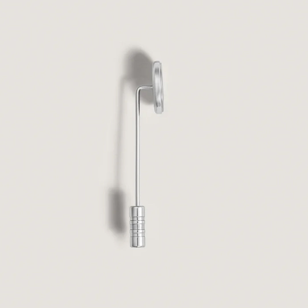 Resembling the Montblanc Great Characters Homage To The Great Gatsby Stainless Steel Lapel Pin, a minimalist silver wall hook features a cylindrical base and curved arm extending to a flat, round top, casting subtle shadows on the light gray wall.
