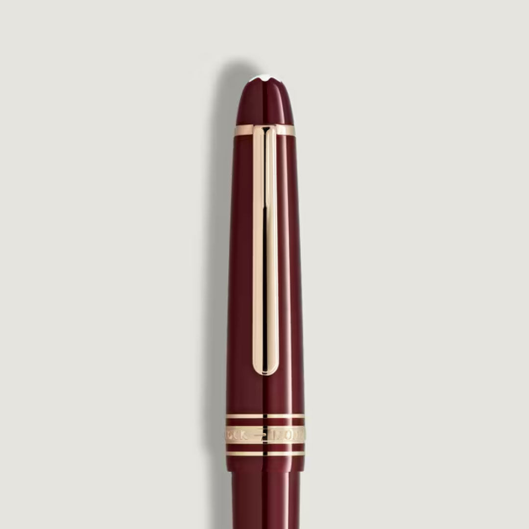 The Montblanc Meisterstuck Burgundy Midsize Ballpoint Pen, featuring gold accents on the cap and clip, stands vertically against a plain light gray background.