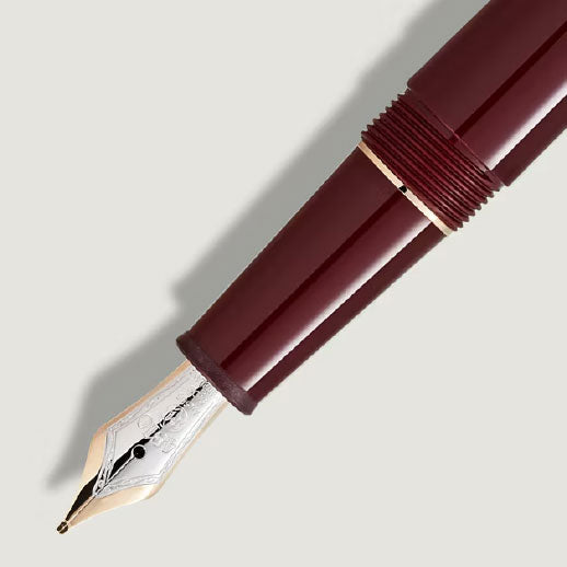 A close-up of the Montblanc Meisterstuck Burgundy Red Classique Fountain Pen, featuring a silver and gold nib, is shown diagonally on a light, neutral background.