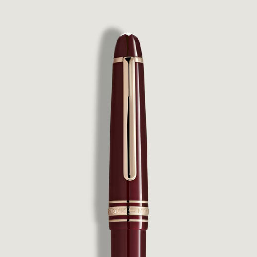 The Montblanc Meisterstuck Burgundy Red Classique Fountain Pen by Montblanc, featuring gold fittings and a gold clip, is displayed vertically against a plain light grey background.