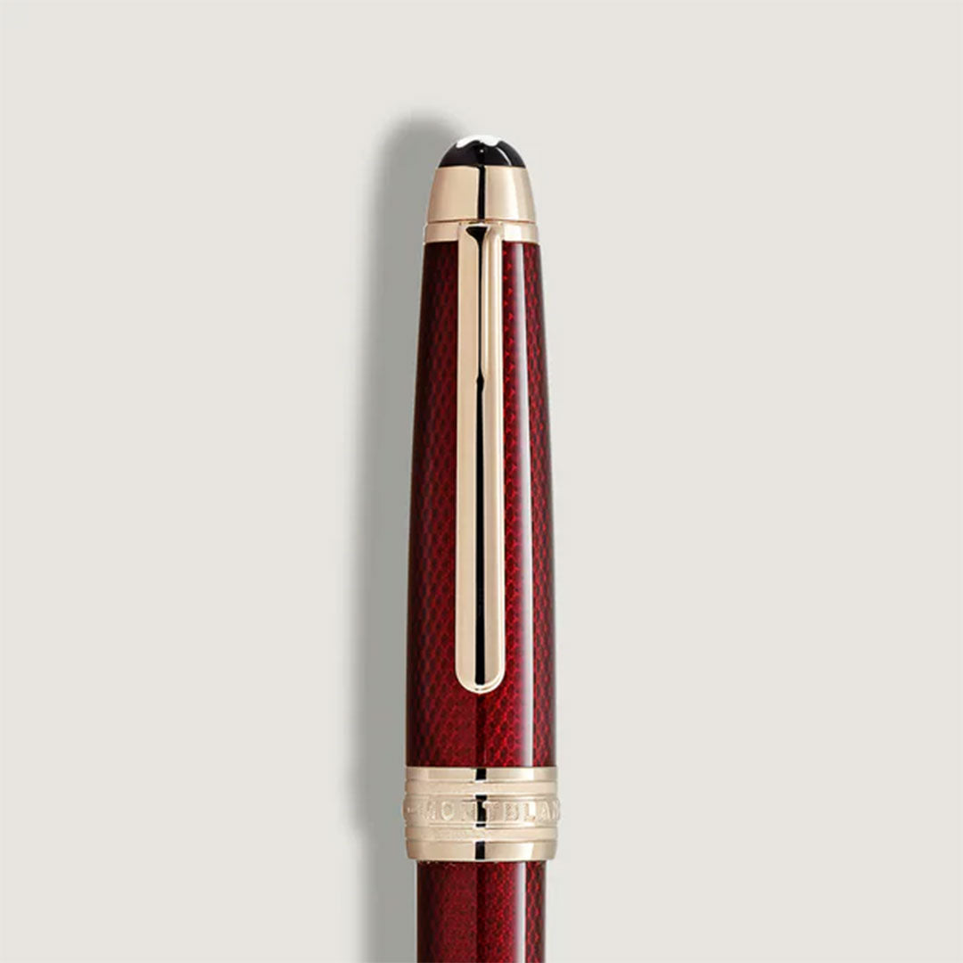 A close-up of the Montblanc Meisterstück Solitaire Golden Hour Ballpoint Pen, a luxury burgundy red lacquer pen with gold-coated fittings and an engraved band near the cap, set against a plain light background.