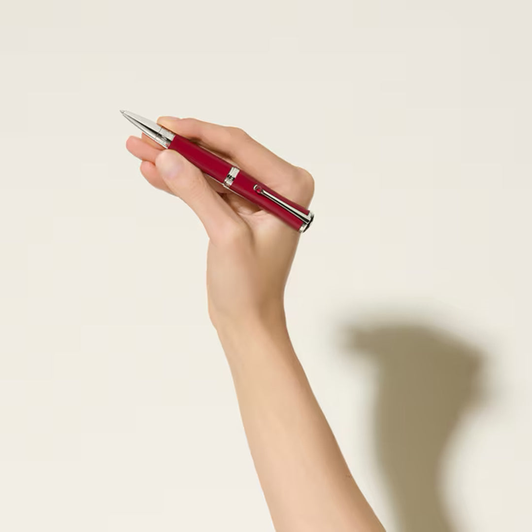 A hand holds the Montblanc Maria Callas Special Edition Ballpoint Pen, ruby-colored and upright, against a plain light background, with a soft shadow cast on the wall.