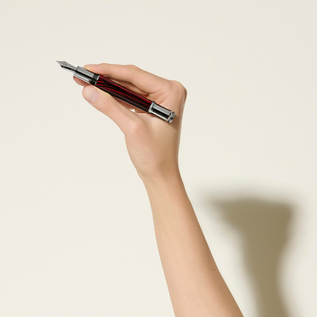 A hand holds the Montblanc Writers Edition Homage to Bram Stoker Limited Edition Fountain Pen, featuring a silver cap and red-and-black striped barrel, against a light background with a soft shadow.