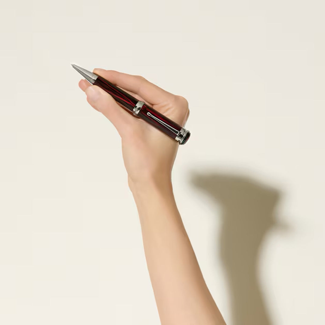 A hand holds the Montblanc Writers Edition Homage to Bram Stoker Limited Edition Ballpoint Pen, featuring black and red stripes, against a plain light background that casts a shadow on the wall.