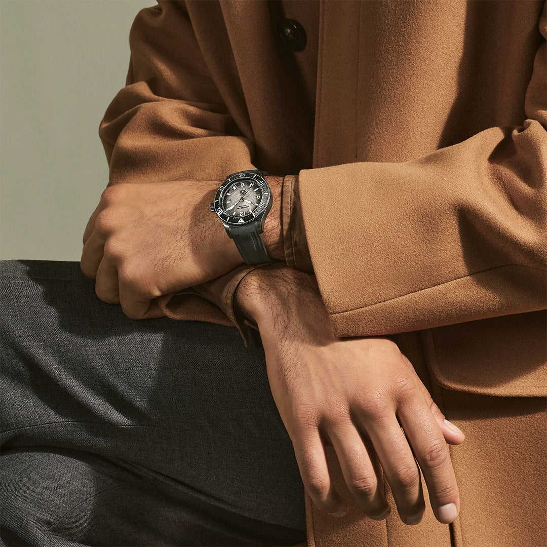 A person in a tan coat and dark gray pants sits with arms crossed, featuring a Montblanc Iced Sea Automatic Date 0 Oxygen 41mm watch on their left wrist. The stylish Montblanc dive watch stands out with its round face and metal accents.