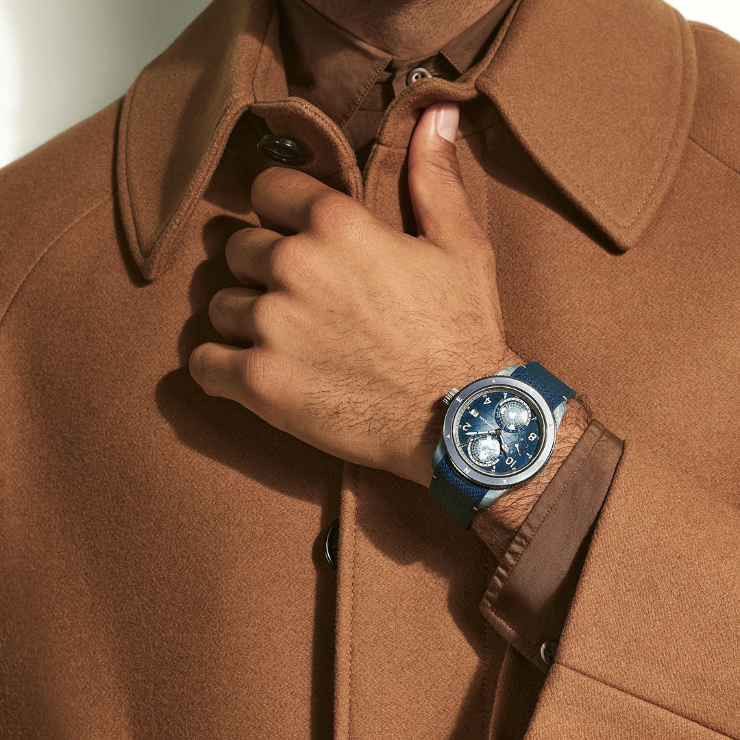 A person wearing a camel coat with a buttoned collar adjusts it near the neck, showcasing a Montblanc 1858 Geosphere 0 Oxygen Mount Vison Automatic 43.5mm Limited Edition watch (986 pcs) on their left wrist.