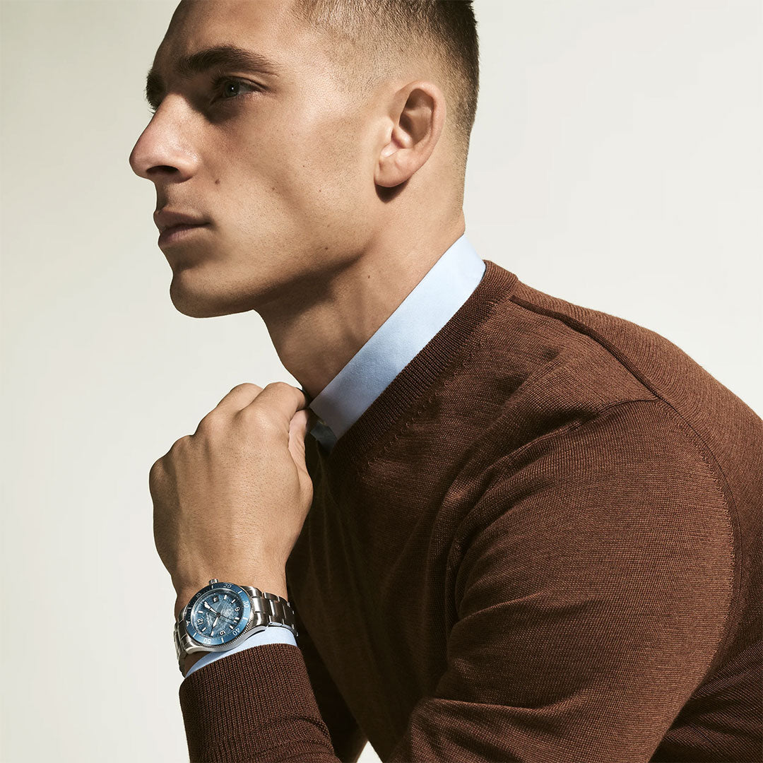 A man in a brown sweater over a light blue collared shirt poses in profile, resting his hand near his chin. He wears the Montblanc Iced Sea Automatic Date 0 Oxygen 38mm Watch with a striking glacier-pattern dial.