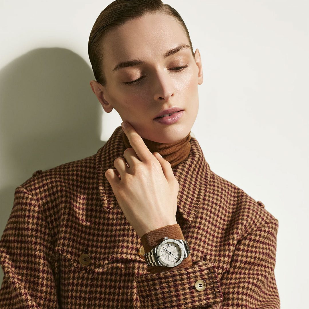A person in a brown houndstooth coat and turtleneck poses with their hand near their chin, displaying the Montblanc Iced Sea Automatic Date 0 Oxygen 38mm Watch with its striking glacier-patterned dial.