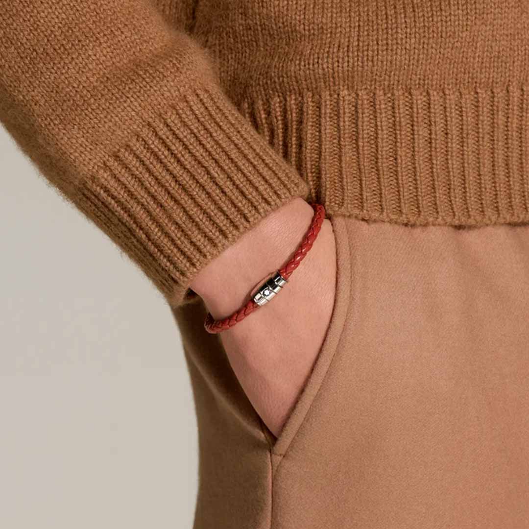 A person wearing a brown sweater and pants has their hand in their pocket. They are wearing the Montblanc Meisterstuck Collection Steel 3-Ring Rust Leather Bracelet, which features a small polished stainless steel clasp. The background is neutral.