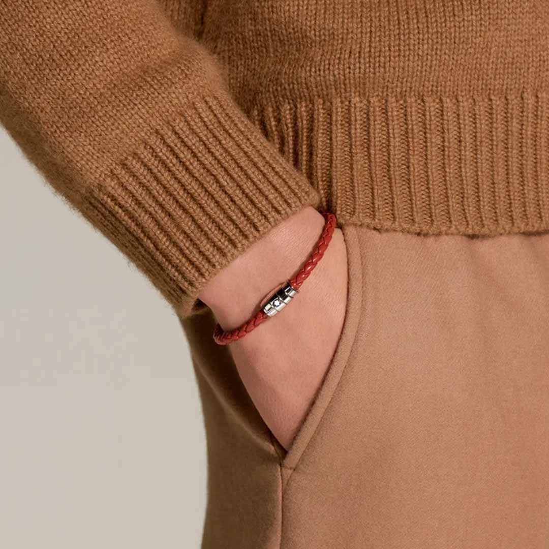 A person wearing a camel-colored sweater and pants has one hand in their pocket. On their wrist, they sport the Montblanc Meisterstuck Collection Steel 3-Ring Rust Leather Bracelet, adding a sleek touch to their look with its polished stainless steel clasp.