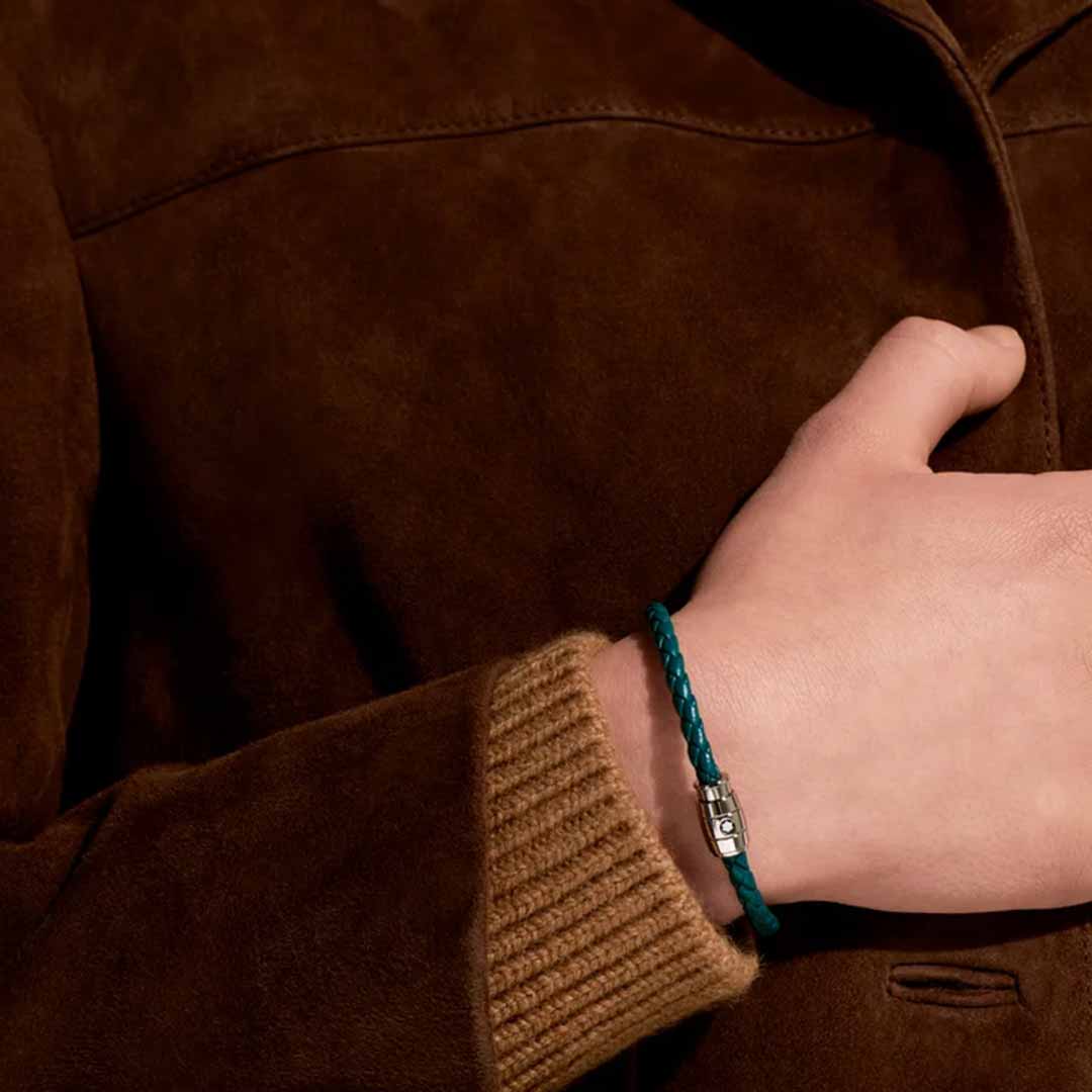 A person dressed in a suede jacket with ribbed sweater cuffs has their hand placed on their chest. On their wrist, they wear the Montblanc Meisterstuck Collection Steel 3-Ring Cyprus Blue Leather Bracelet from Montblanc.