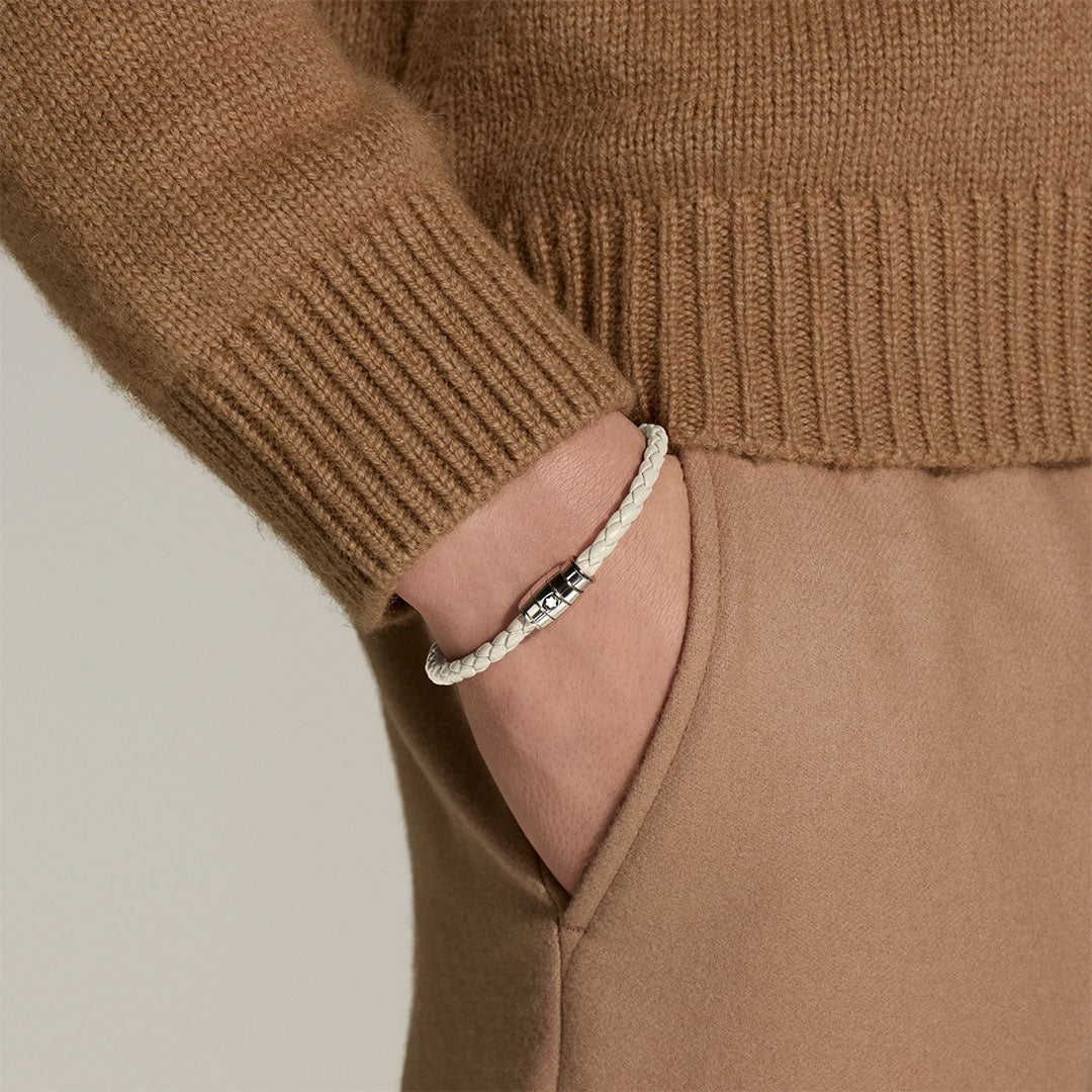 A person in a brown sweater and pants has their hand in their pocket, displaying the Montblanc Meisterstück 3 Rings Cloudy Beige Leather Bracelet with a signature emblem and magnetic clasp on their wrist.