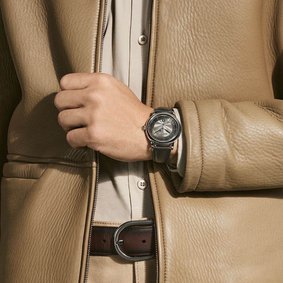 A person in a tan leather jacket and beige shirt is shown from the neck down, resting their left hand on their chest and wearing a Montblanc Star Legacy Automatic Date 42mm Watch by Montblanc with an anthracite dial.