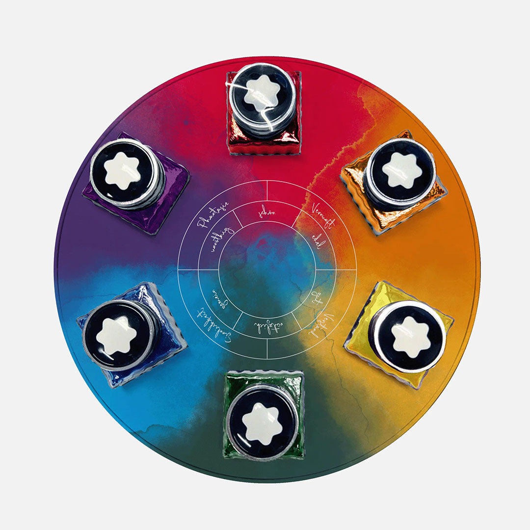 The Montblanc Writers Edition Homage to Johann Wolfgang von Goethe 6x30ml Ink Bottles Set displays six vibrant bottles arranged on a multicolored surface around a script-filled white circle, reflecting the spirit of Goethe’s legacy.